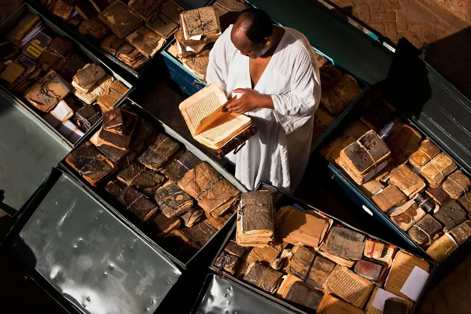 Un homme vêtu de blanc consulte un manuscrit ancien parmi des caisses de manuscrits